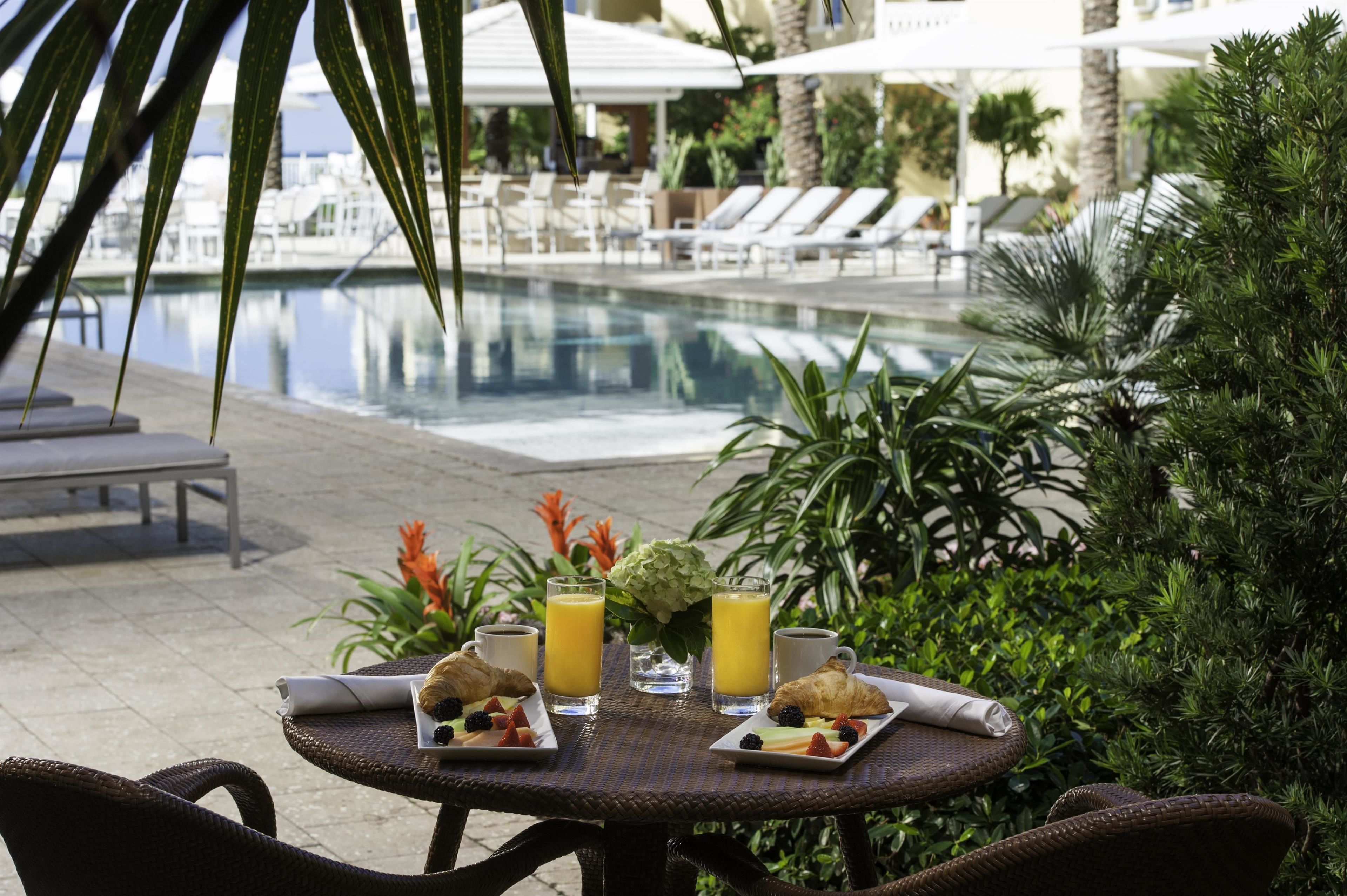 Une table à manger dressée avec des aliments et des boissons pour le petit-déjeuner, donnant sur un espace piscine équipé de chaises longues et de parasols.