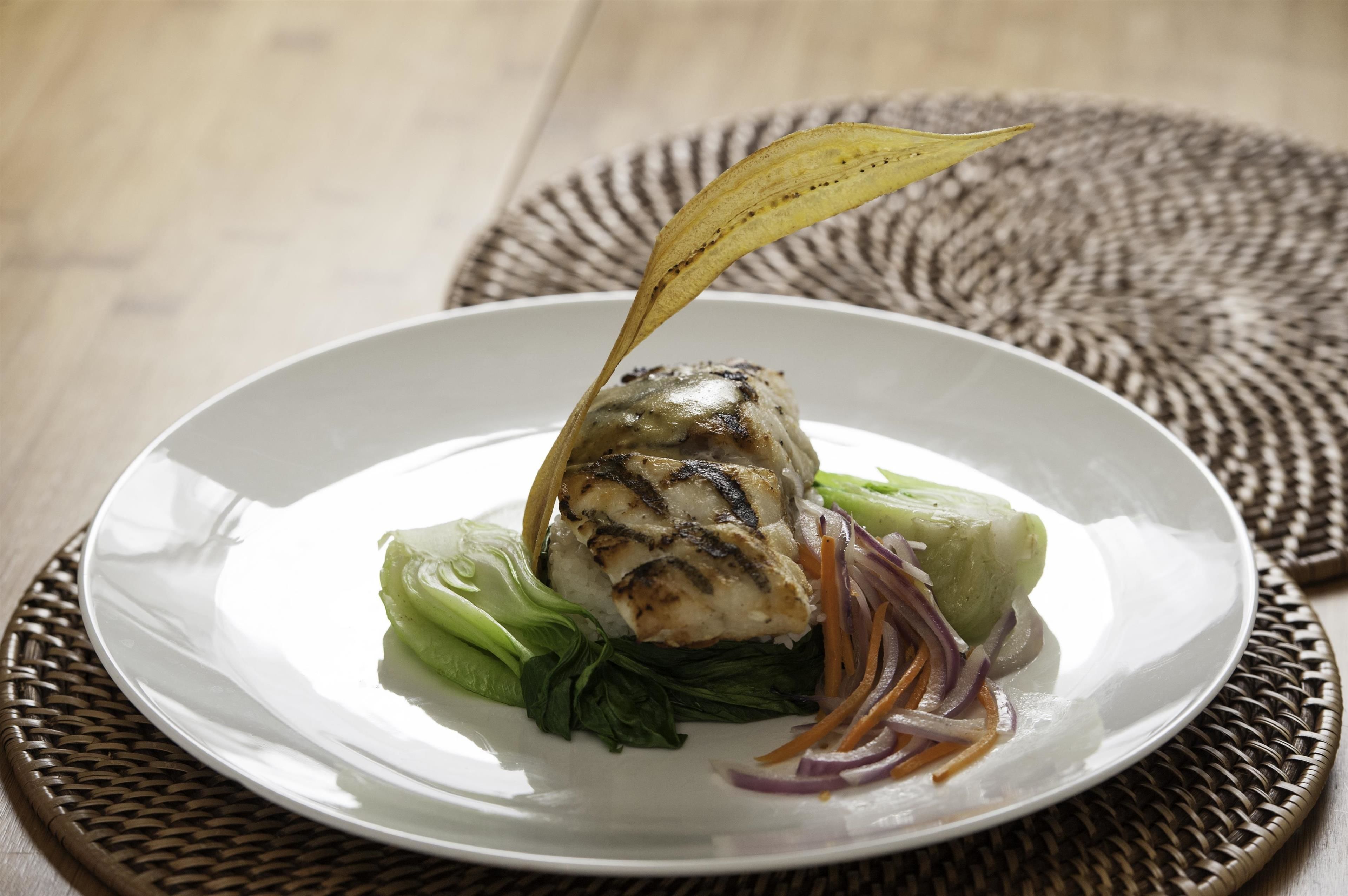 Um prato branco com frango grelhado, vegetais verdes e uma folha decorativa sobre um jogo americano tecido.