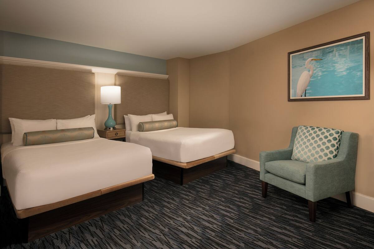 A hotel room with two beds, a nightstand with a lamp, a chair, and a framed picture on the wall.