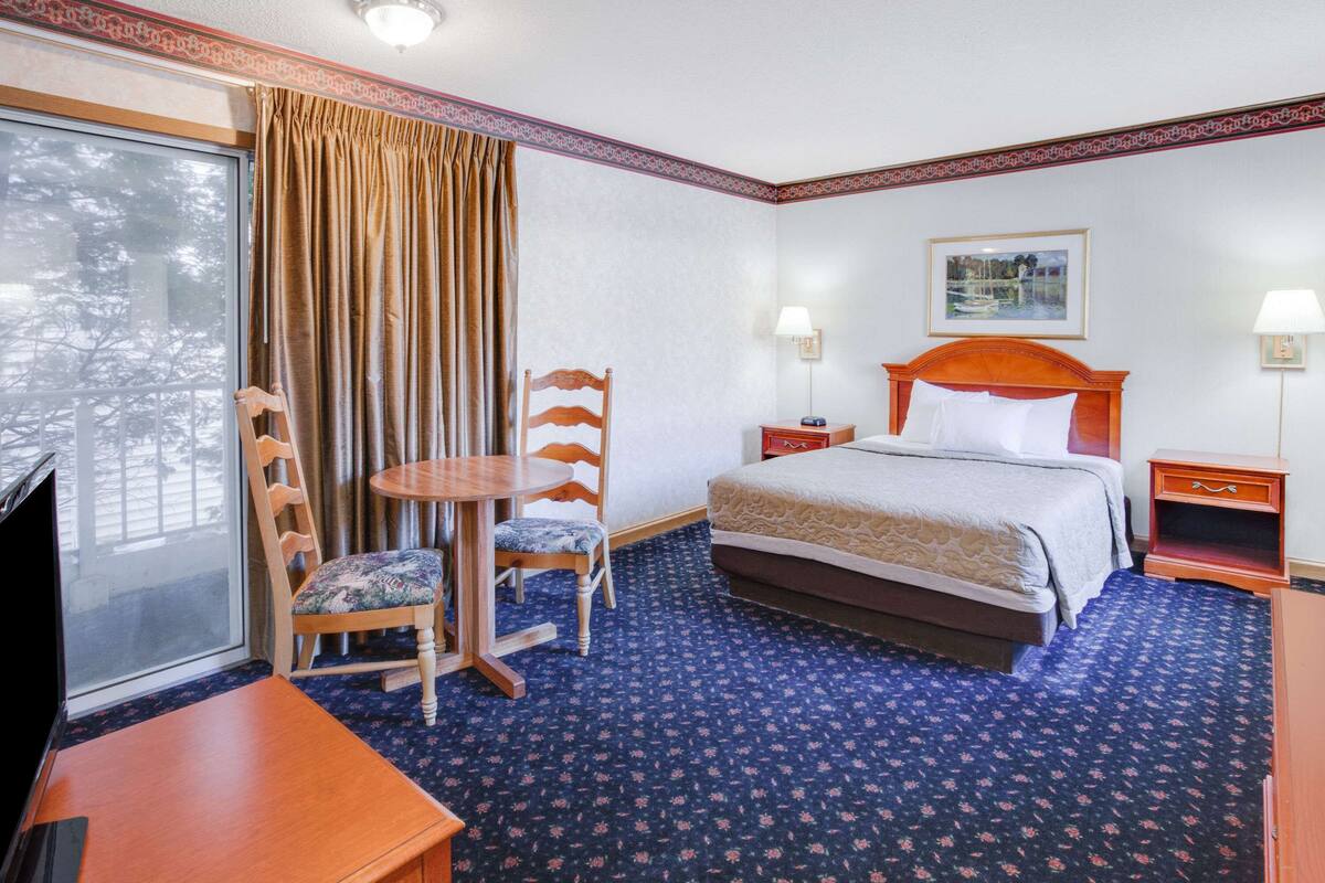 Ein Hotelzimmer mit großem Fenster, einem Bett mit Holz Kopfteil, einem kleinen Tisch mit zwei Stühlen, einem Nachttisch mit Lampe und einem gerahmten Bild an der Wand.