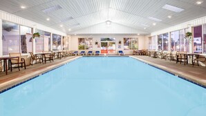 An indoor swimming pool with clear blue water, surrounded by wooden deck chairs and tables, and large windows on both sides. - Super 8 by Wyndham Mackinaw City/Beachfront Area (Mackinaw City)