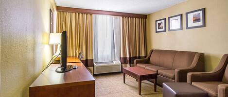 Una habitación de hotel con un escritorio, un sofá de cuero marrón, una mesa de centro y un ventanal con cortinas. Hay tres cuadros en la pared.