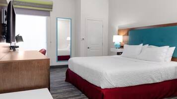 A hotel room with a bed, a desk, a chair, a nightstand with a lamp, and a window with blinds.