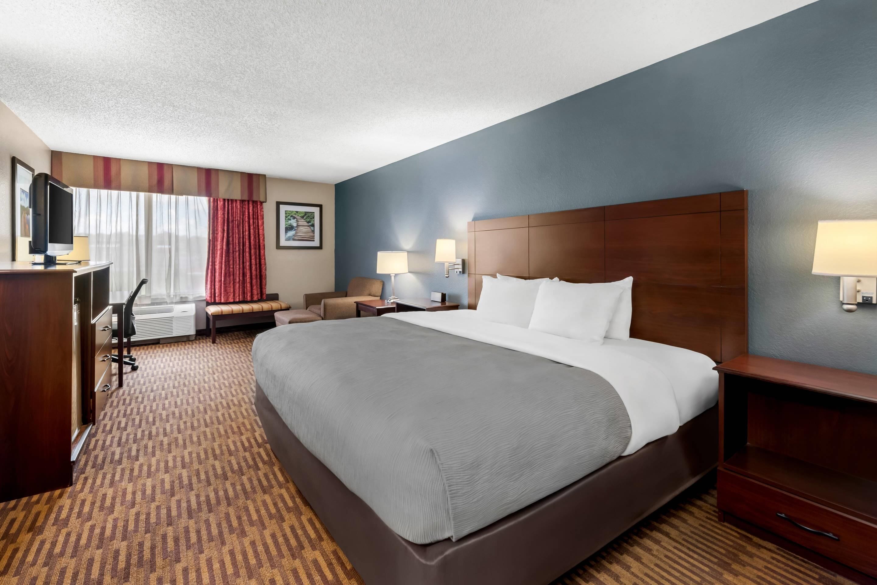 A hotel room with a large bed, a desk with a television, a chair, a bench, two bedside lamps, and a carpeted floor.