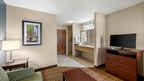 A hotel room with a sofa, a TV, a desk, a lamp, a framed picture, a bathroom with a sink and mirror, and a wooden door.