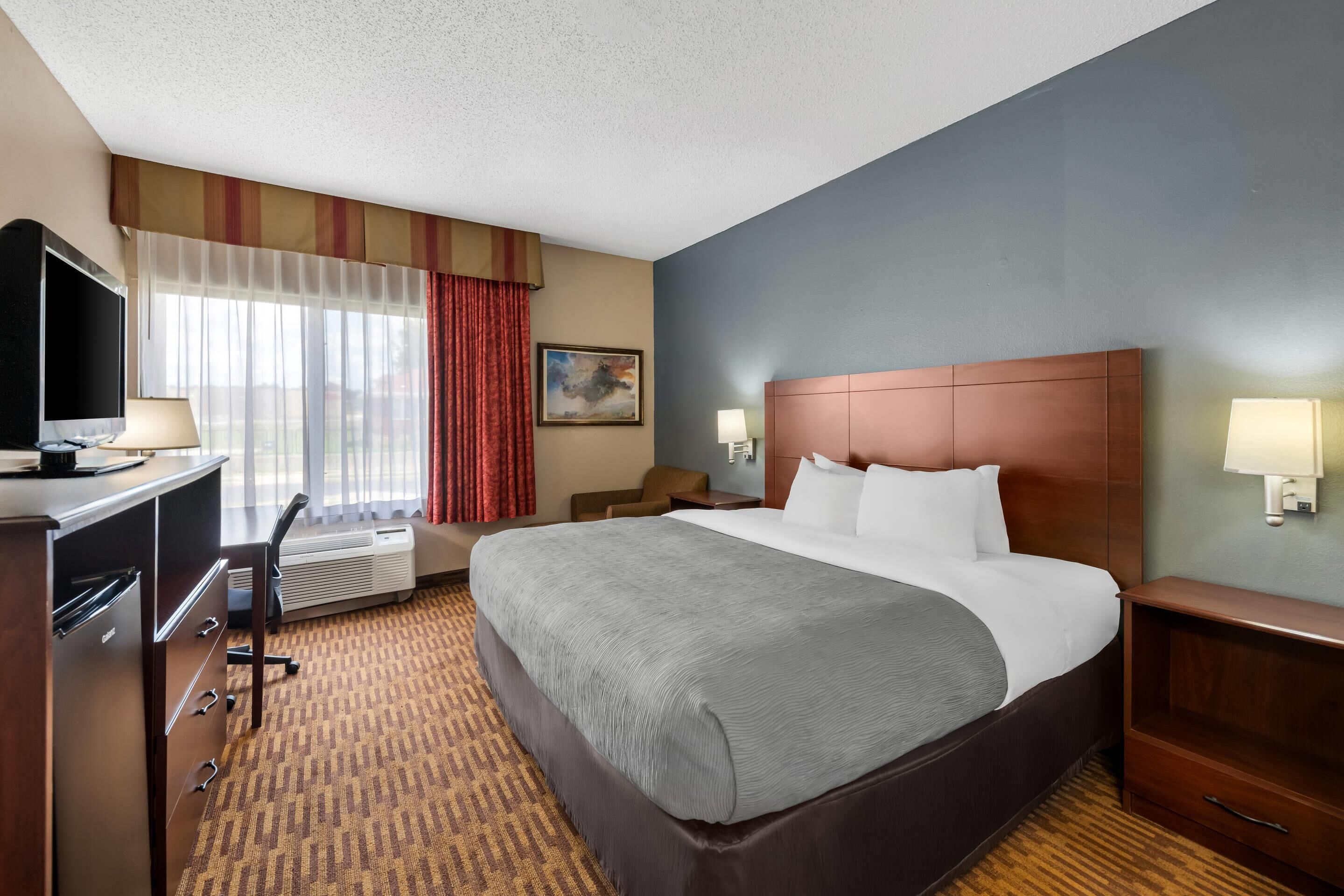A hotel room with a large bed, a desk with a computer, a TV, a window with curtains, and a painting on the wall.
