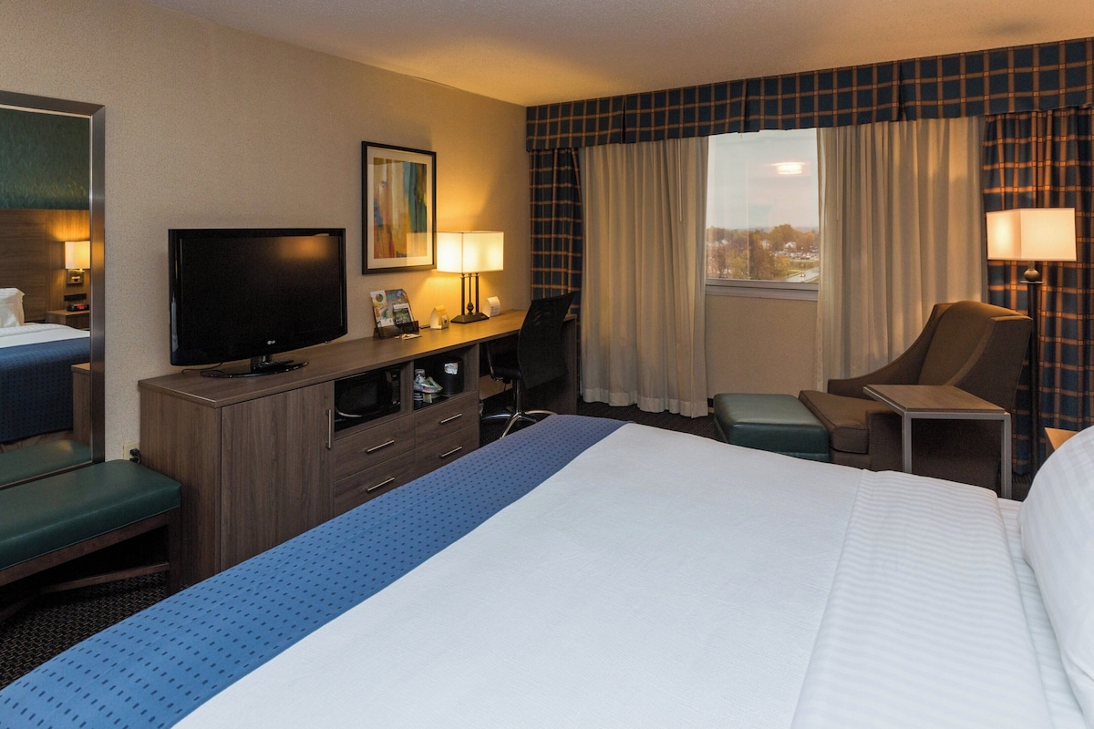Ein Hotelzimmer mit einem Bett, einem Schreibtisch mit Computer, einem Fernseher, einem Sessel und einem Fenster mit Vorhängen.