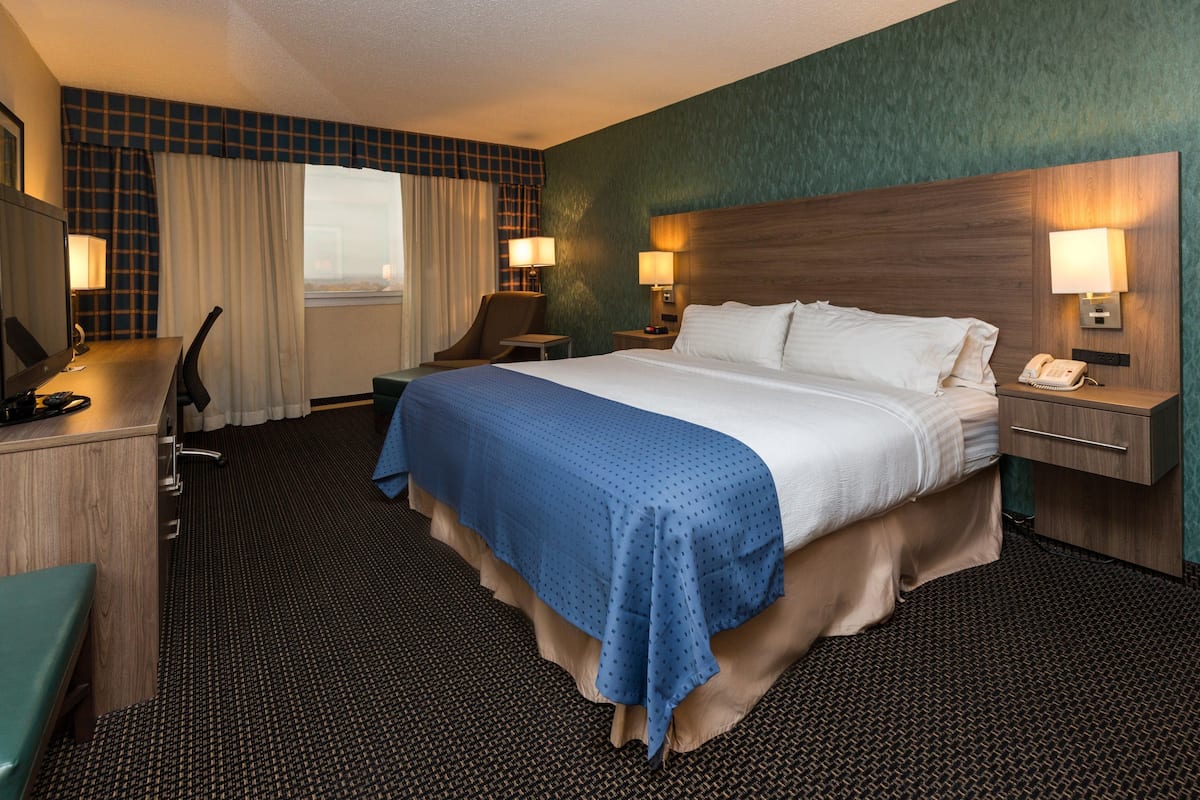 A hotel room with a large bed, a desk, a chair, a nightstand with a phone, and a window with curtains.