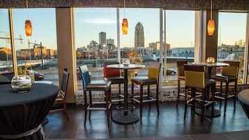 An indoor seating area with tables and chairs, large windows with a cityscape view, and hanging pendant lights.