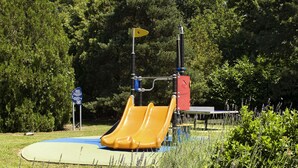 Un parque infantil con un tobogán amarillo y una zona de juegos azul, rodeado de hierba alta y árboles.