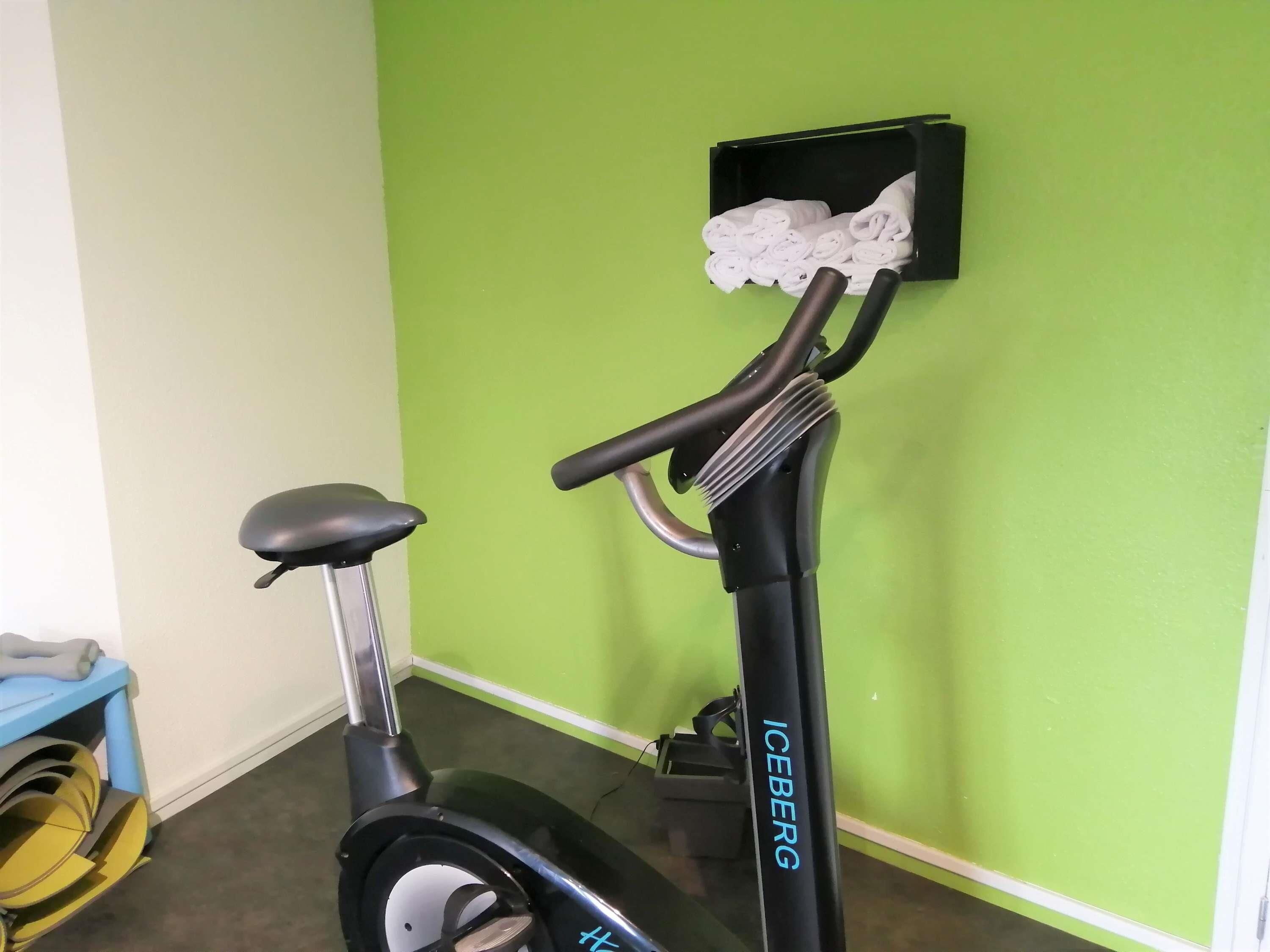 Una bicicleta estática con un compartimento que contiene toallas dobladas, un carro azul con ruedas amarillas y una pared verde.