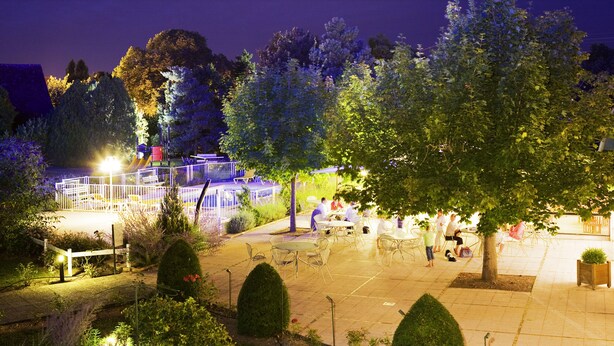 Un patio al aire libre con mesas y sillas, rodeado de árboles y un parque infantil al fondo.