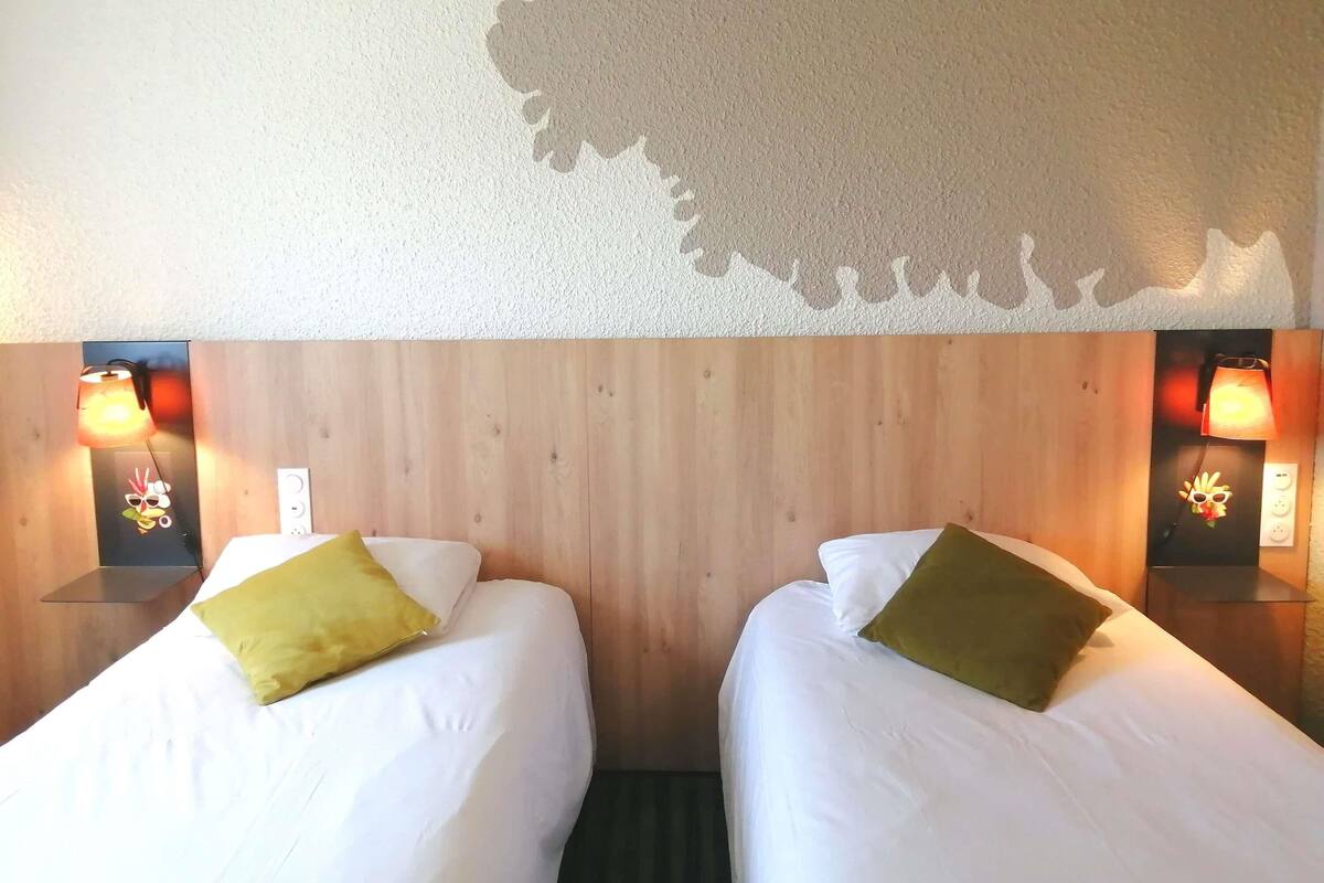 Une chambre d’hôtel avec deux lits, chacun équipé d’un oreiller, d’une tête de lit en bois et d’une lampe murale dont l’abat-jour présente un motif décoratif.