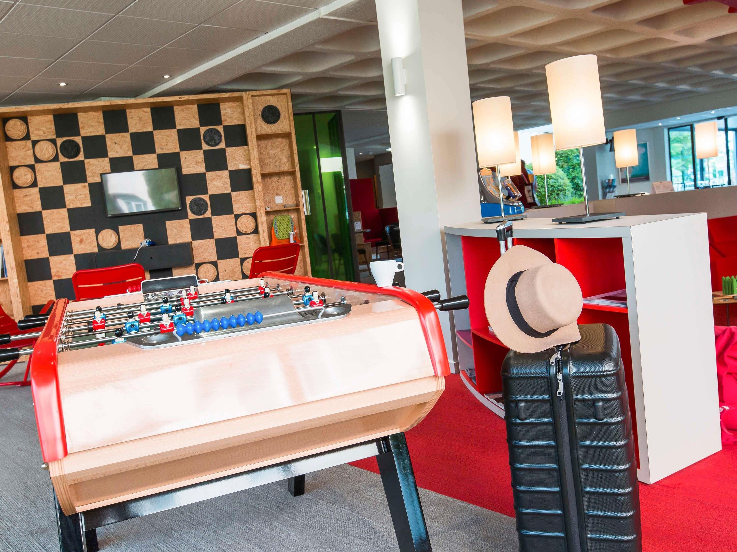 A foosball table, a red sofa, and a hat on a suitcase in a modern room with a checkered wall and a red carpet.