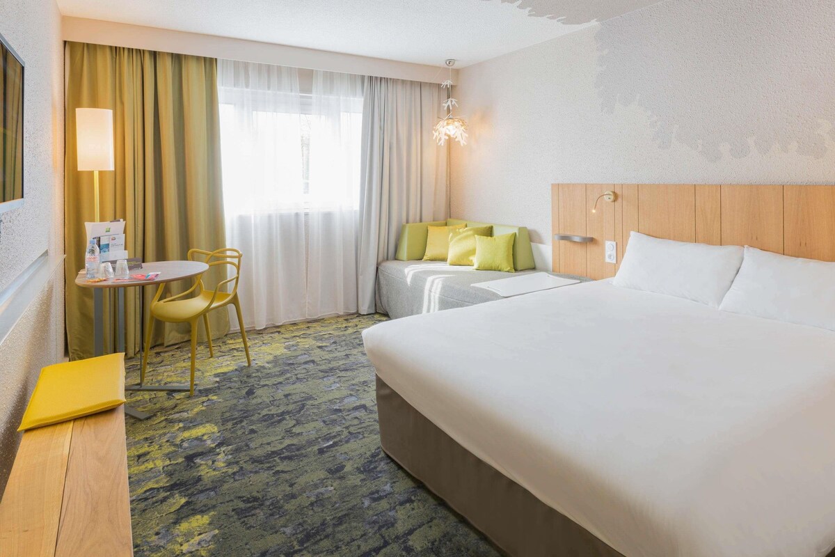 A hotel room with a large bed, a desk with a chair, a small table with a chair, and a window with curtains.