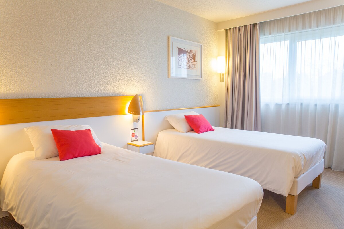 A hotel room with two beds, a nightstand with a lamp, a framed picture on the wall, and a window with curtains.