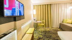 Habitación de hotel con televisión montada en la pared, una mesita redonda con una silla, un banco, una cama y un ventanal con cortinas.