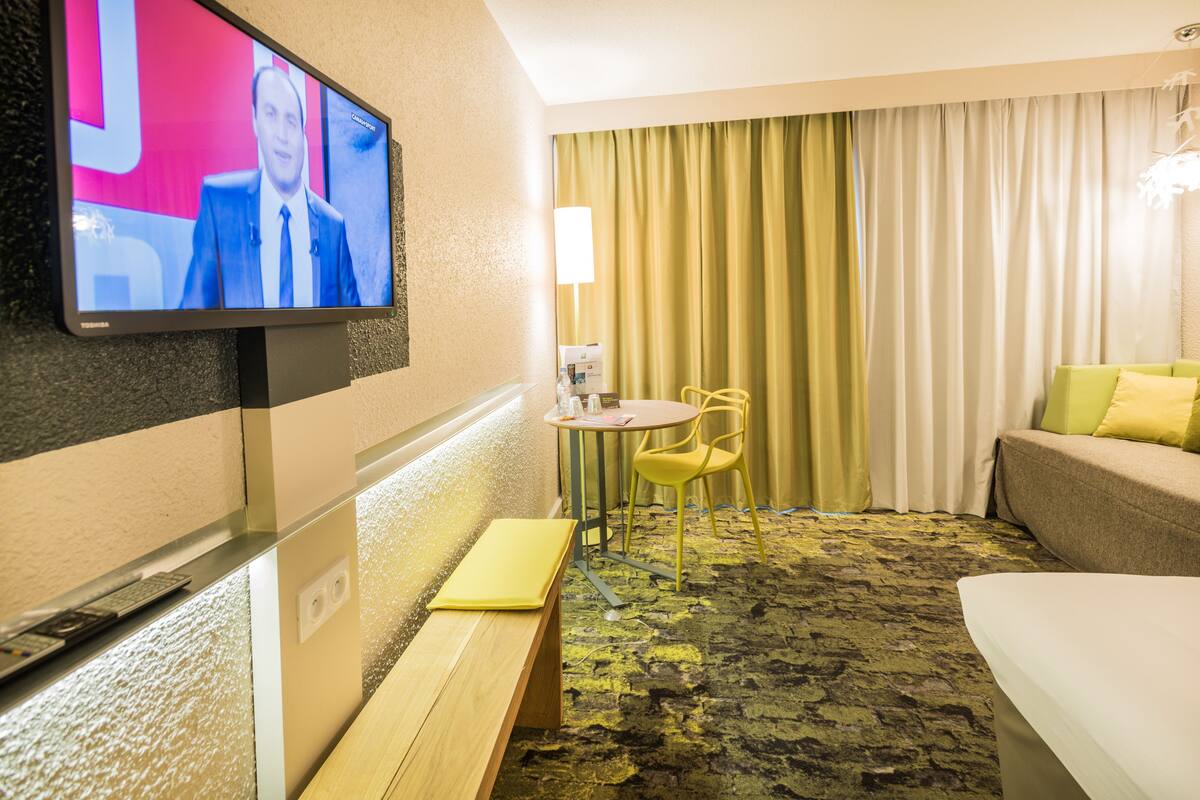 Une chambre d’hôtel équipée d’un téléviseur fixé au mur, d’une petite table ronde avec une chaise, d’un banc, d’un lit et d’une grande fenêtre avec des rideaux.