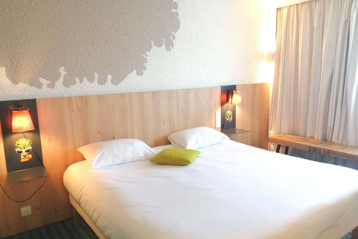 Une chambre d’hôtel avec un grand lit, deux lampes de chevet, une fenêtre avec des rideaux et une étagère fixée au mur.