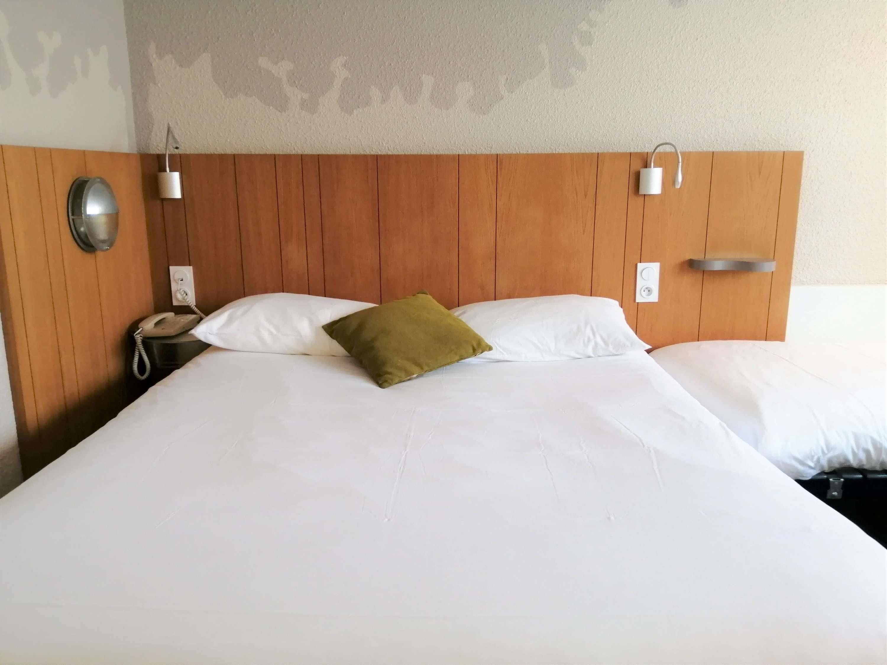 Habitación de hotel con cabecera de madera, dos lámparas de noche, un teléfono y una cama con ropa de cama blanca y almohada verde.