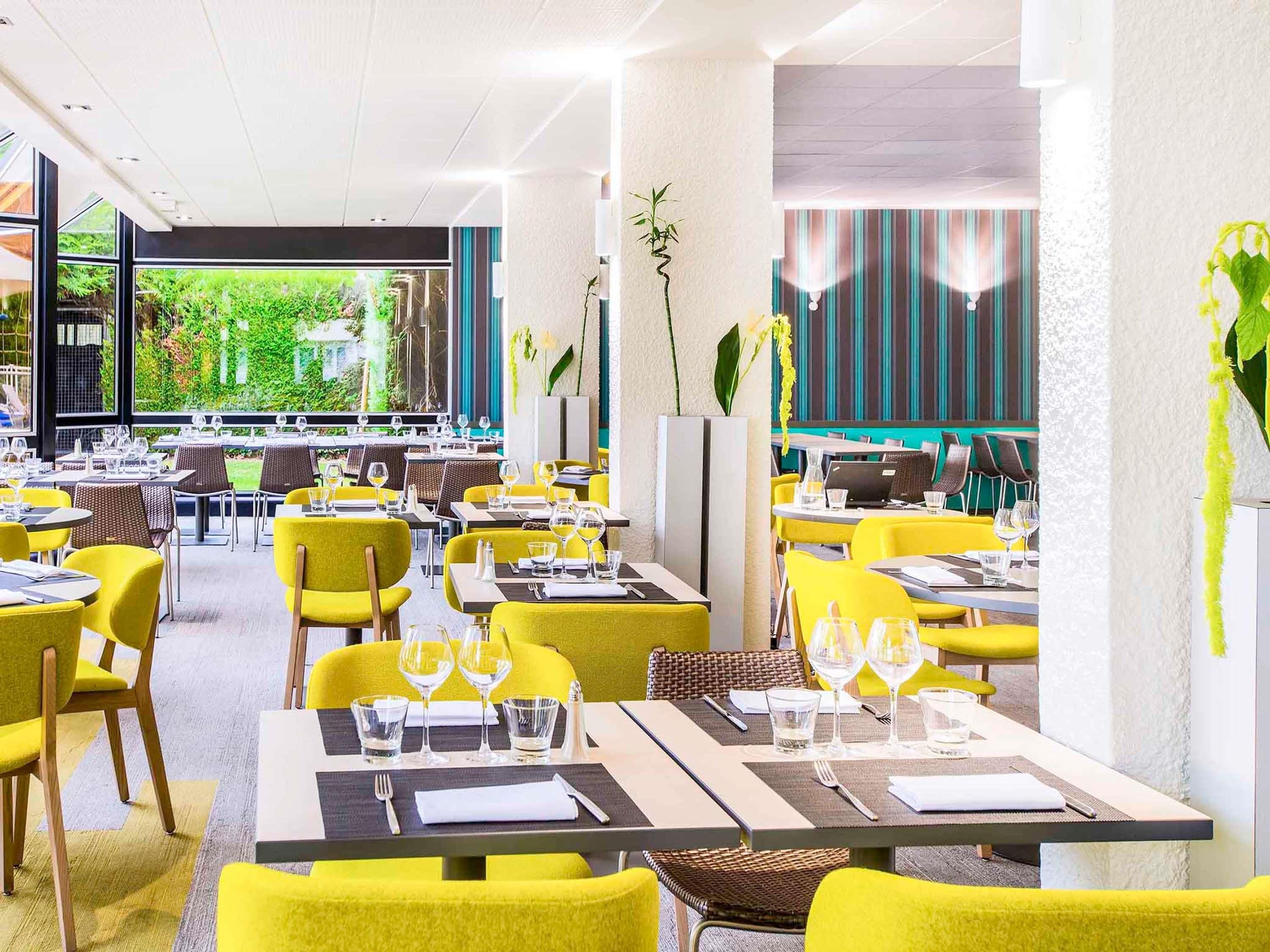A modern restaurant with yellow chairs and tables, large windows offering a view of greenery, and a mix of indoor and outdoor seating.
