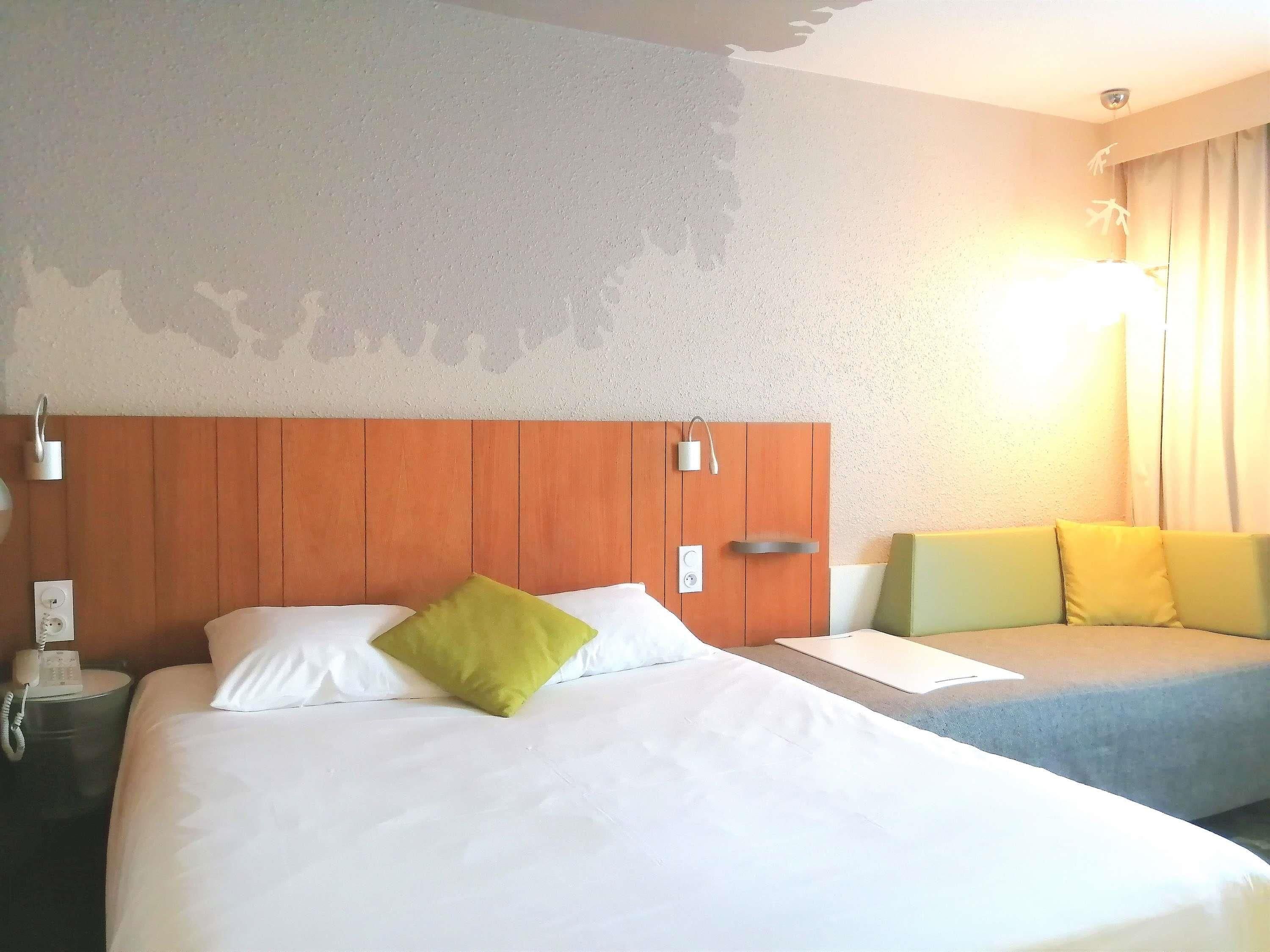 A hotel room with a wooden headboard, a bed with white linens and a green pillow, a bedside table with a phone, and a nightstand.
