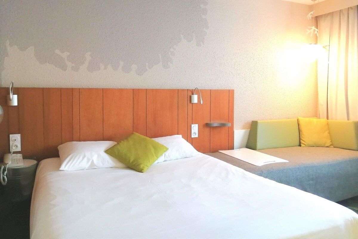 Hotellihuone, jossa on puinen sängynpääty, valkoisilla lakanoiden ja vihreän tyynyn koristeltu sänky, yöpöytä, jossa on puhelin, ja yöpöytäkaappi.
