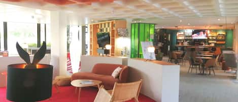 Un lobby moderno de hotel con alfombra roja, un sofá, una mesa y una zona de bar con sillas y una pantalla de televisión.