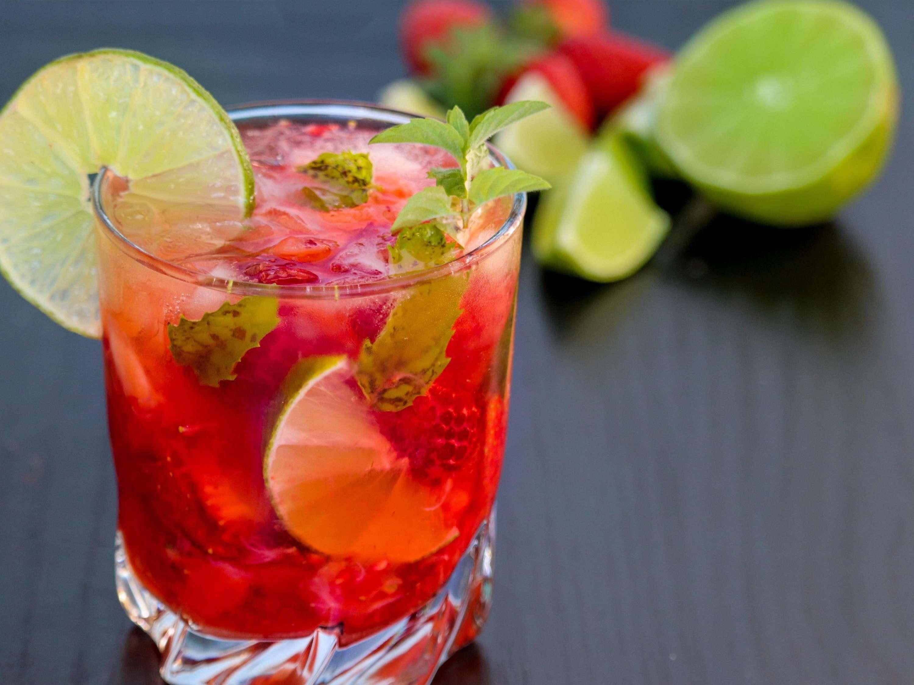 A glass of red beverage with lime slices and mint leaves, with a dark background and a blurred image of limes and strawberries in the background.
