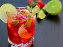 Un vaso de bebida roja con rodajas de lima y hojas de menta, sobre un fondo oscuro y una imagen borrosa de limones y fresas al fondo.