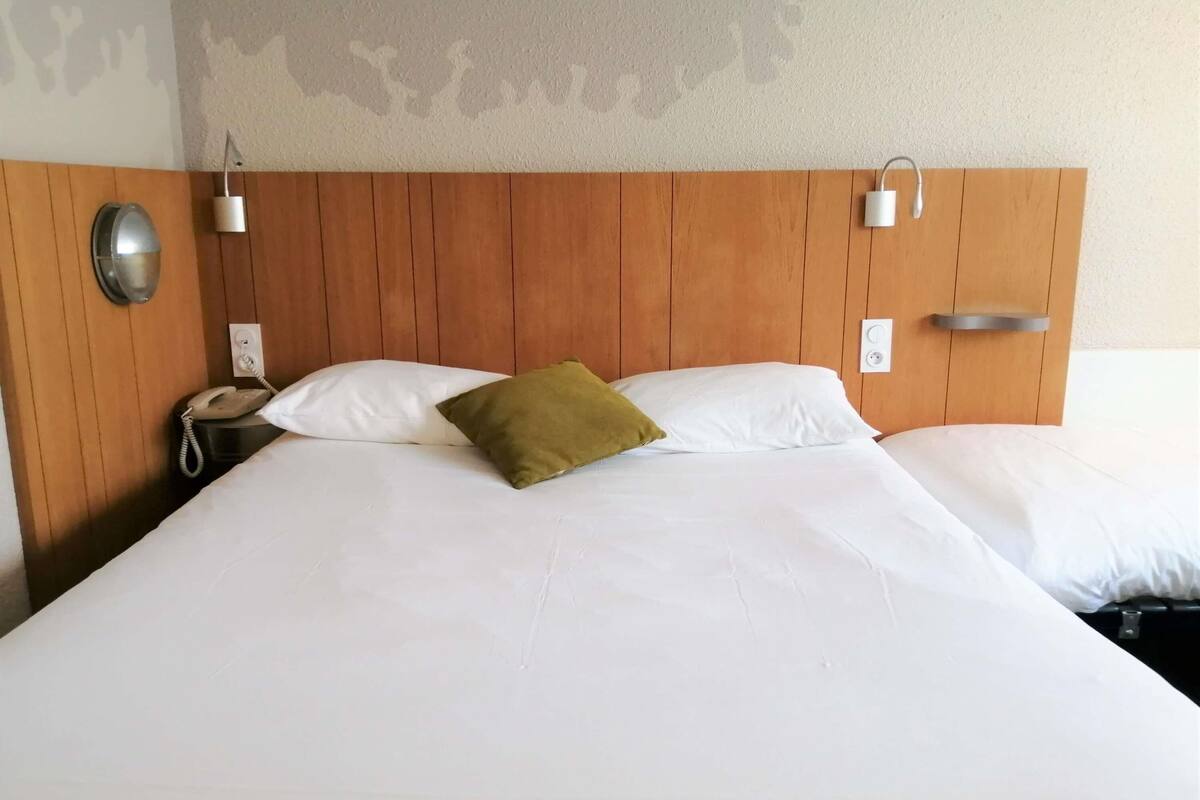 Une chambre d’hôtel avec une tête de lit en bois, deux lampes de chevet, un téléphone et un lit recouvert de draps blancs et agrémenté d’un oreiller vert.