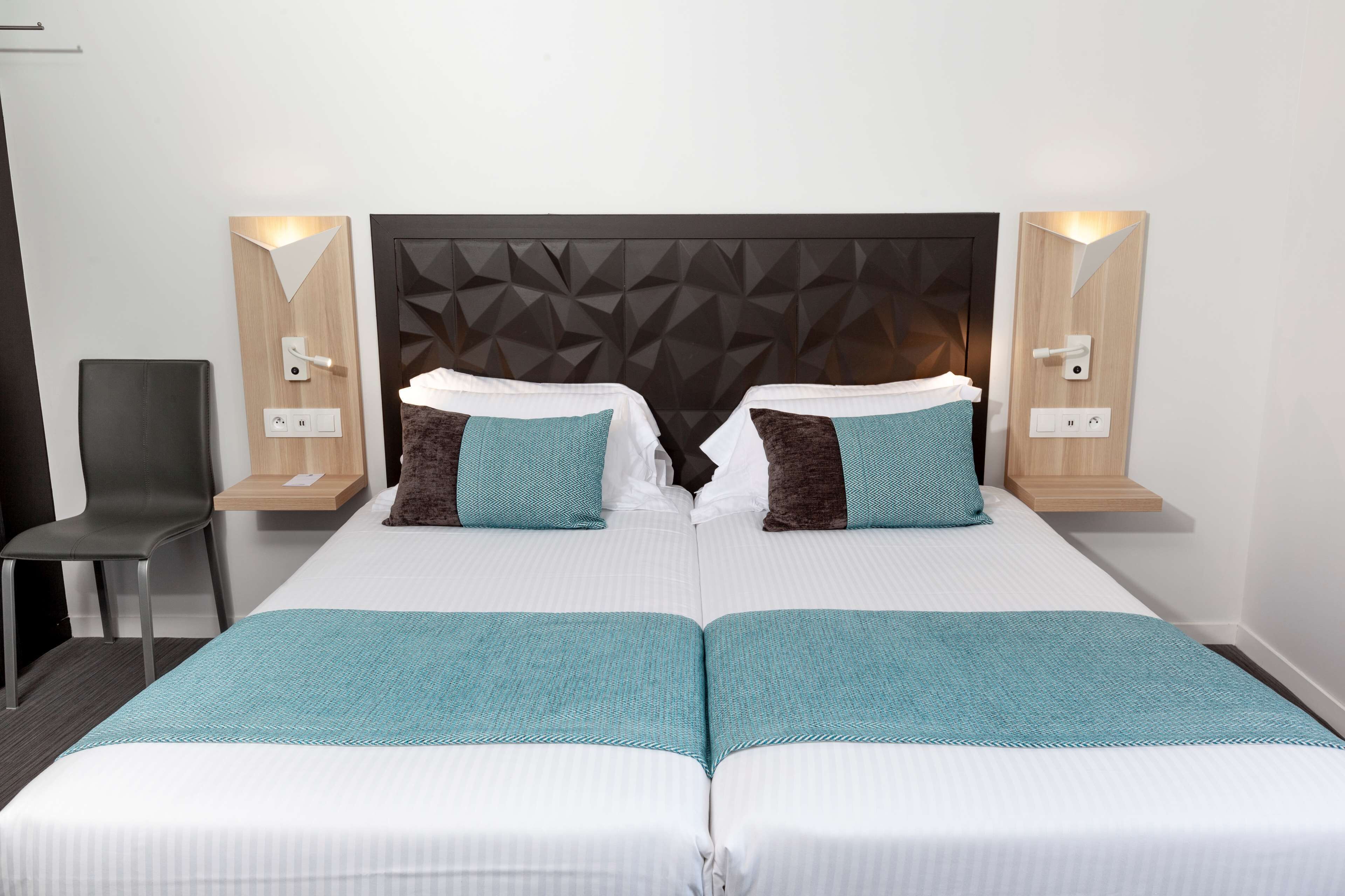 A double bed with a geometric headboard, two bedside tables with lamps, a chair, and a teal throw blanket.