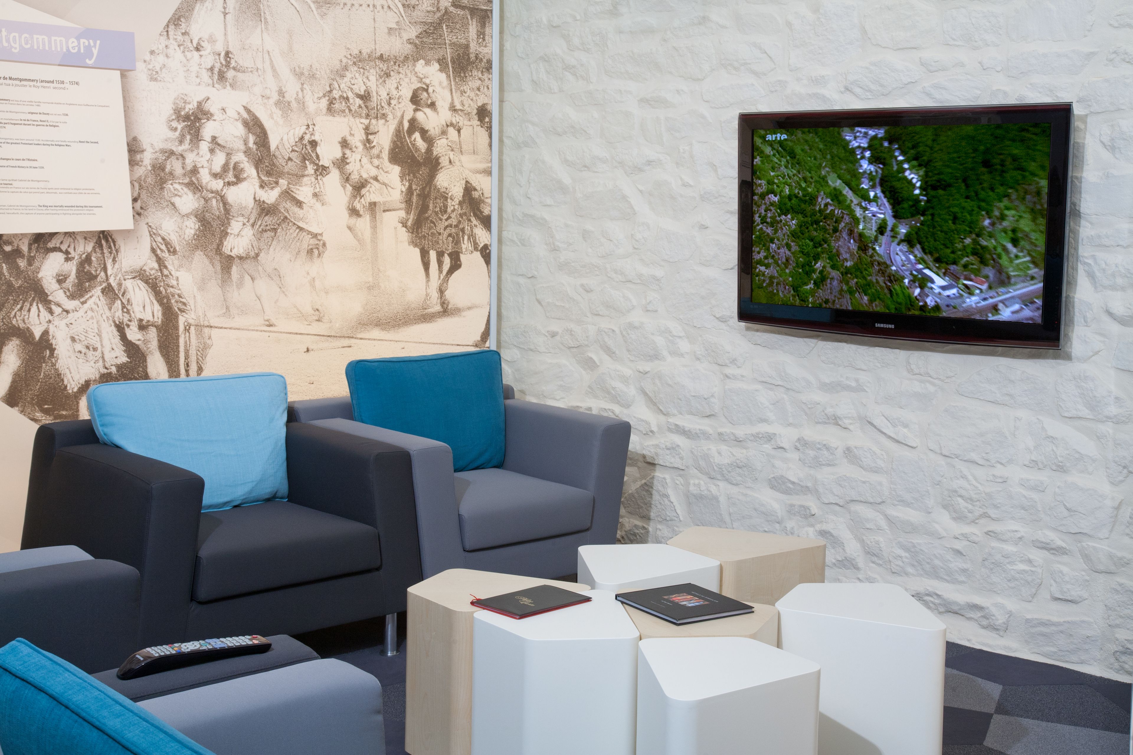 Un salon moderne avec deux canapés gris, un téléviseur à écran plat fixé sur un mur en pierre blanche et une télécommande posée sur une table basse.