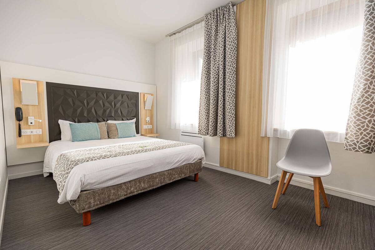 A modern hotel room with a large bed, a chair, and a window with patterned curtains.