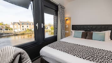 A hotel room with a large window, a bed with patterned bedding, a headboard with geometric design, and a view of buildings across a canal.
