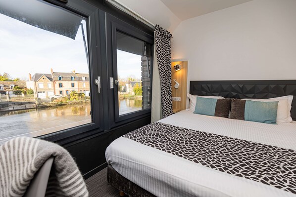 A hotel room with a large window, a bed with patterned bedding, a headboard with geometric design, and a view of buildings across a canal.