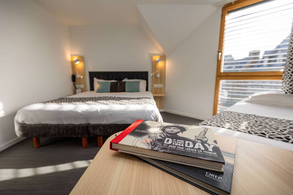 A modern hotel room with a neatly made bed, a bedside table with a book, and a window with blinds.