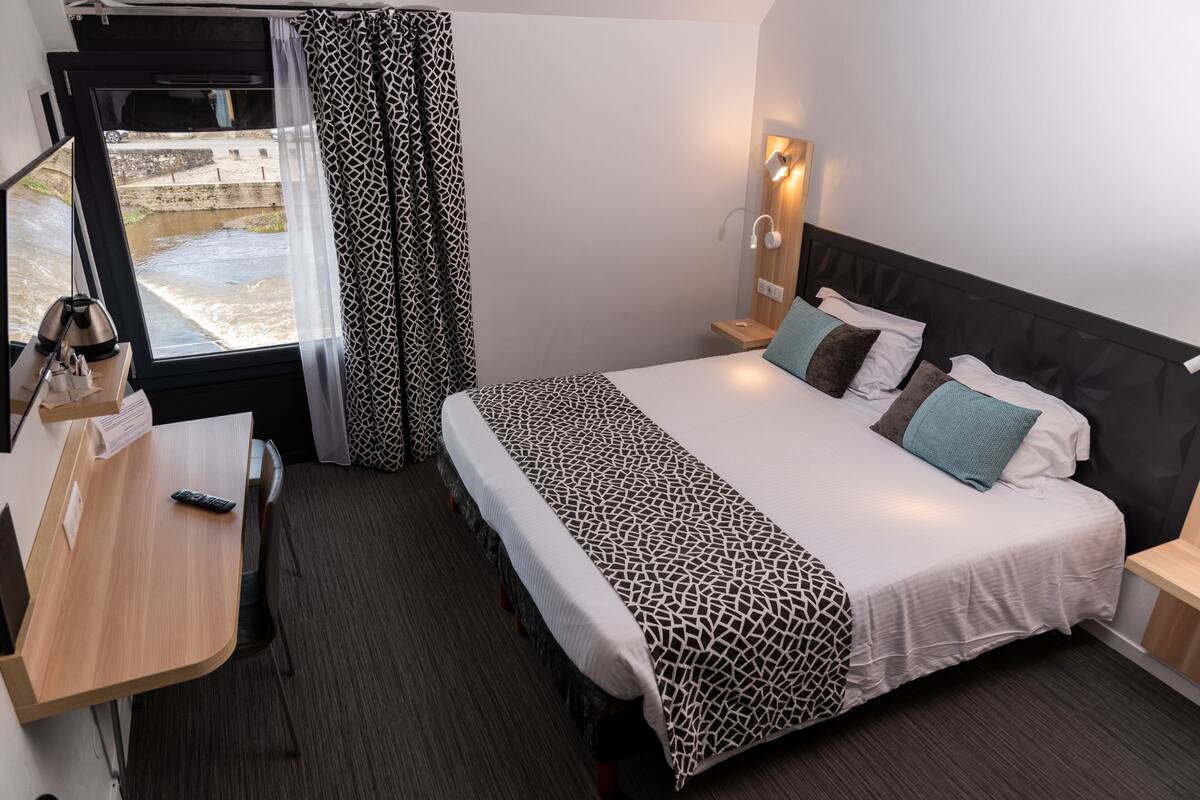 A hotel room with a bed, bedside table, lamp, and a window with curtains.