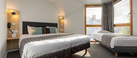 A modern hotel room with two beds, a large window with blinds, and a small desk with a lamp.
