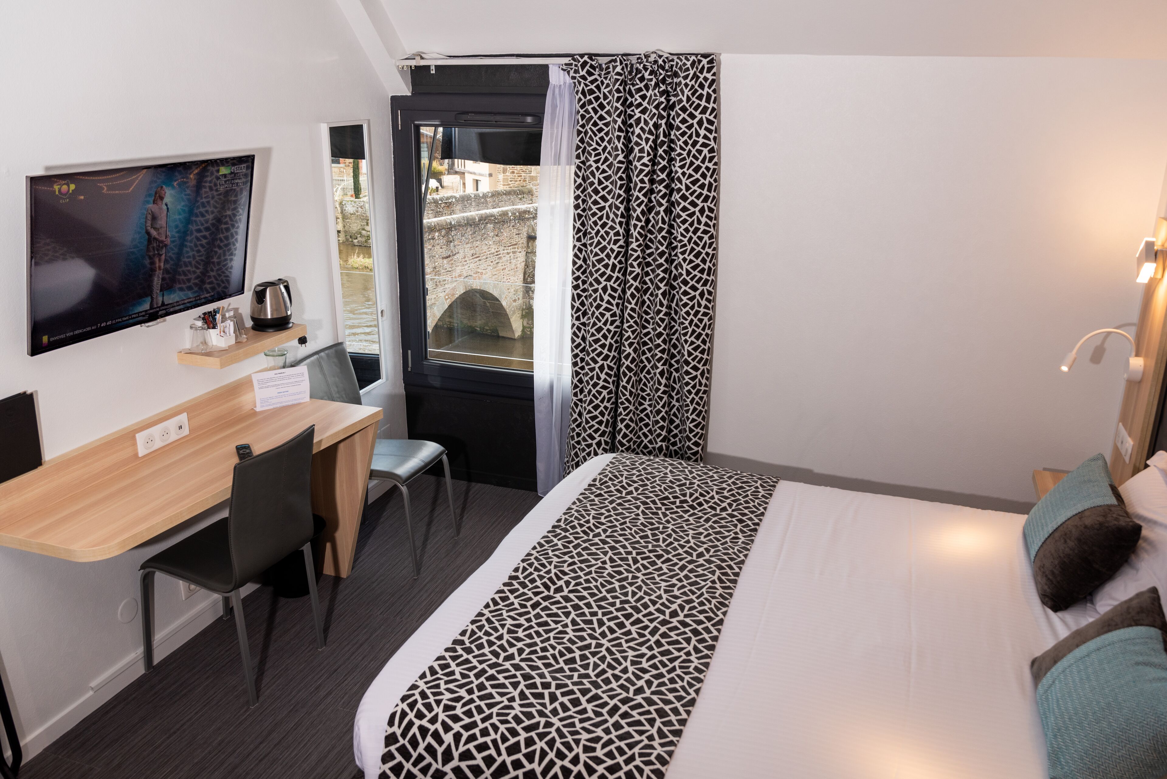 A hotel room with a bed, a desk with a chair, a television, and a window with curtains.