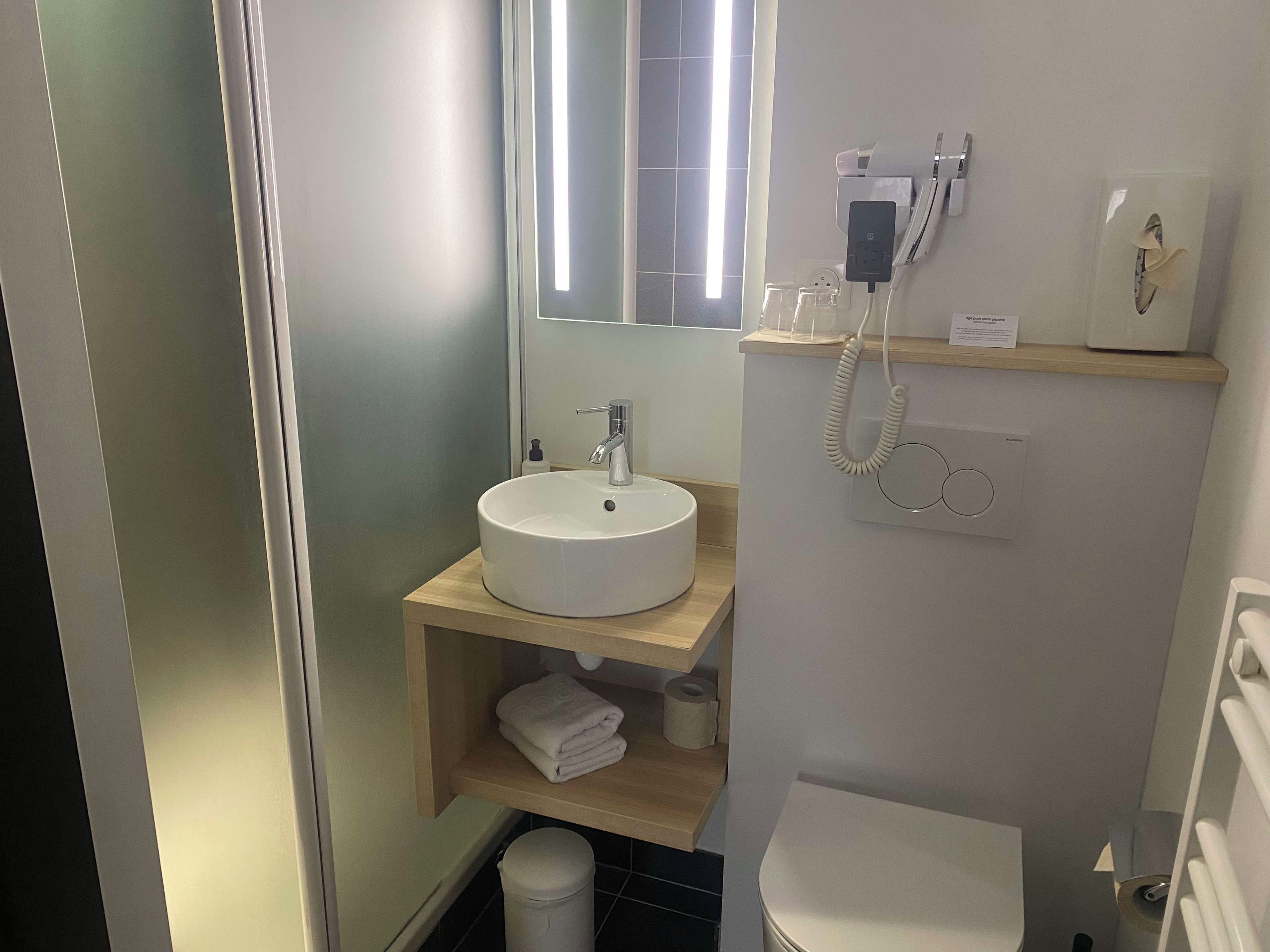 A bathroom with a white sink, mirror, and toilet. There is a towel rack and a shelf with toiletries.