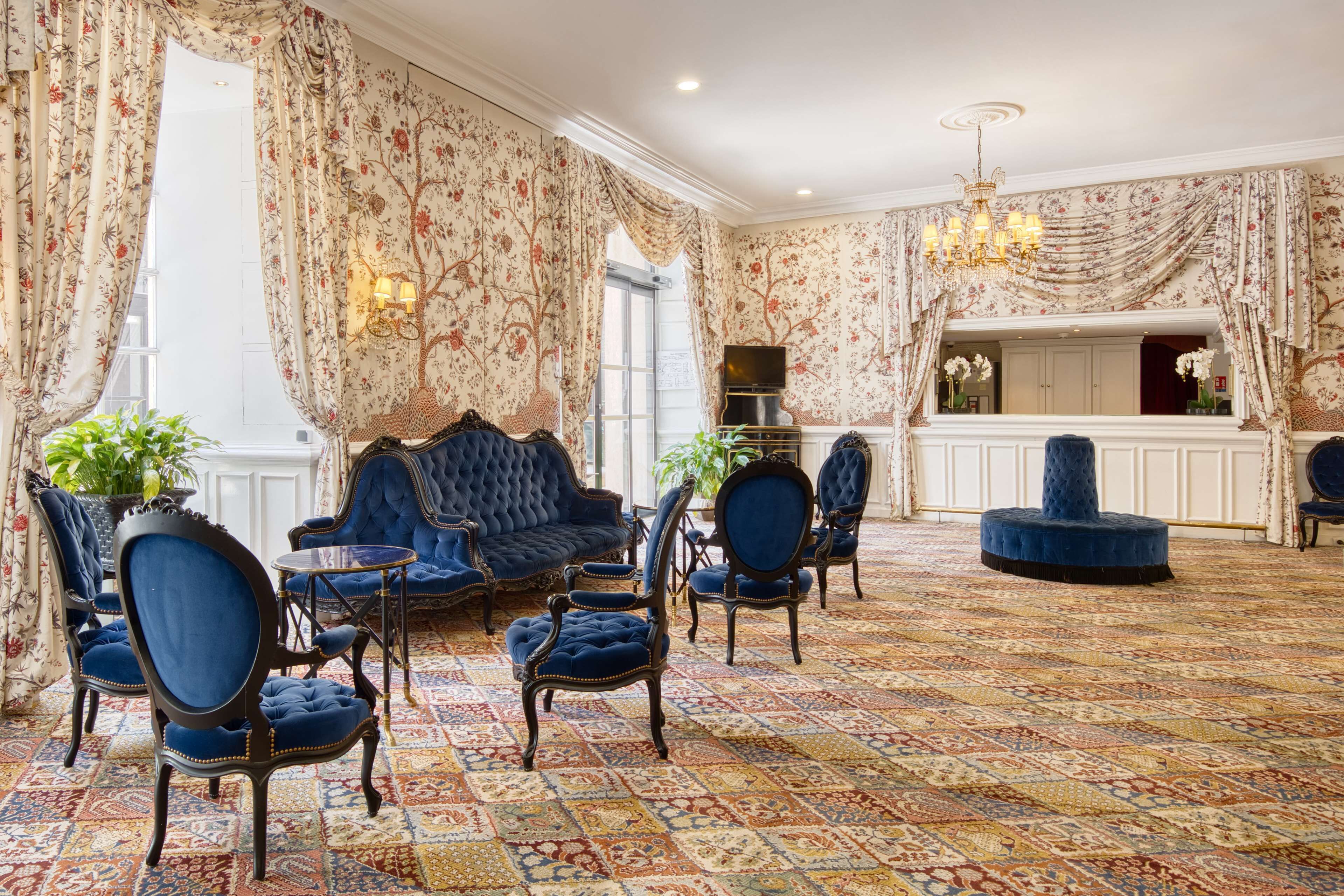 A room with a patterned carpet, blue upholstered chairs, a sofa, and a chandelier.