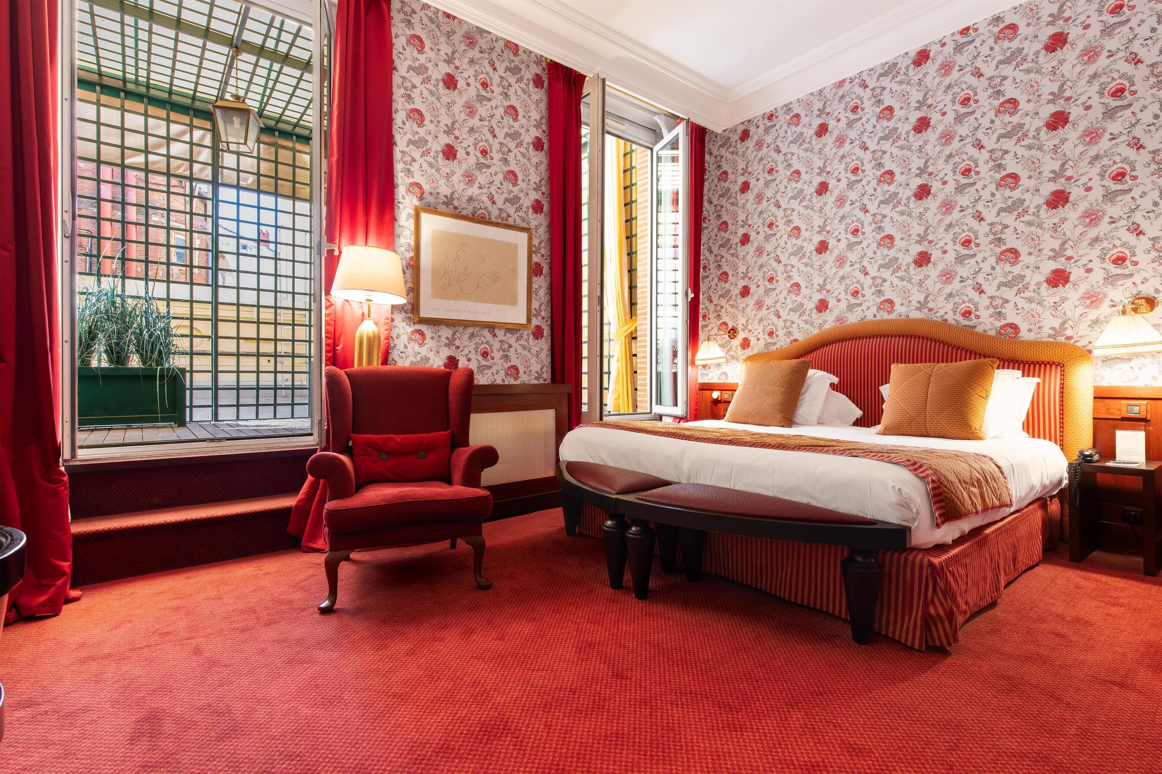 A hotel room with a large bed, a red armchair, a small table, and a window with red curtains.