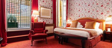 A hotel room with a large bed, a red armchair, a small table, and a window with red curtains.