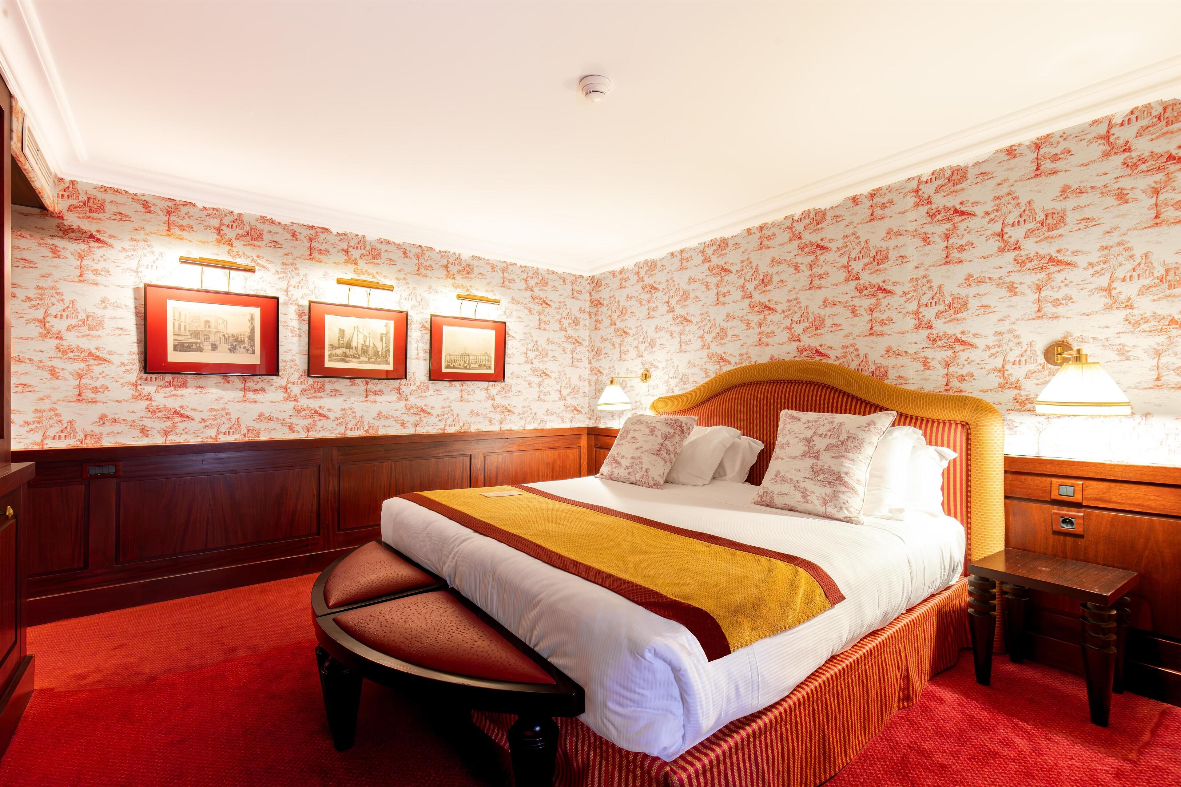 A hotel room with a large bed, a wooden nightstand, a desk, and a chair. There are three framed pictures on the wall.