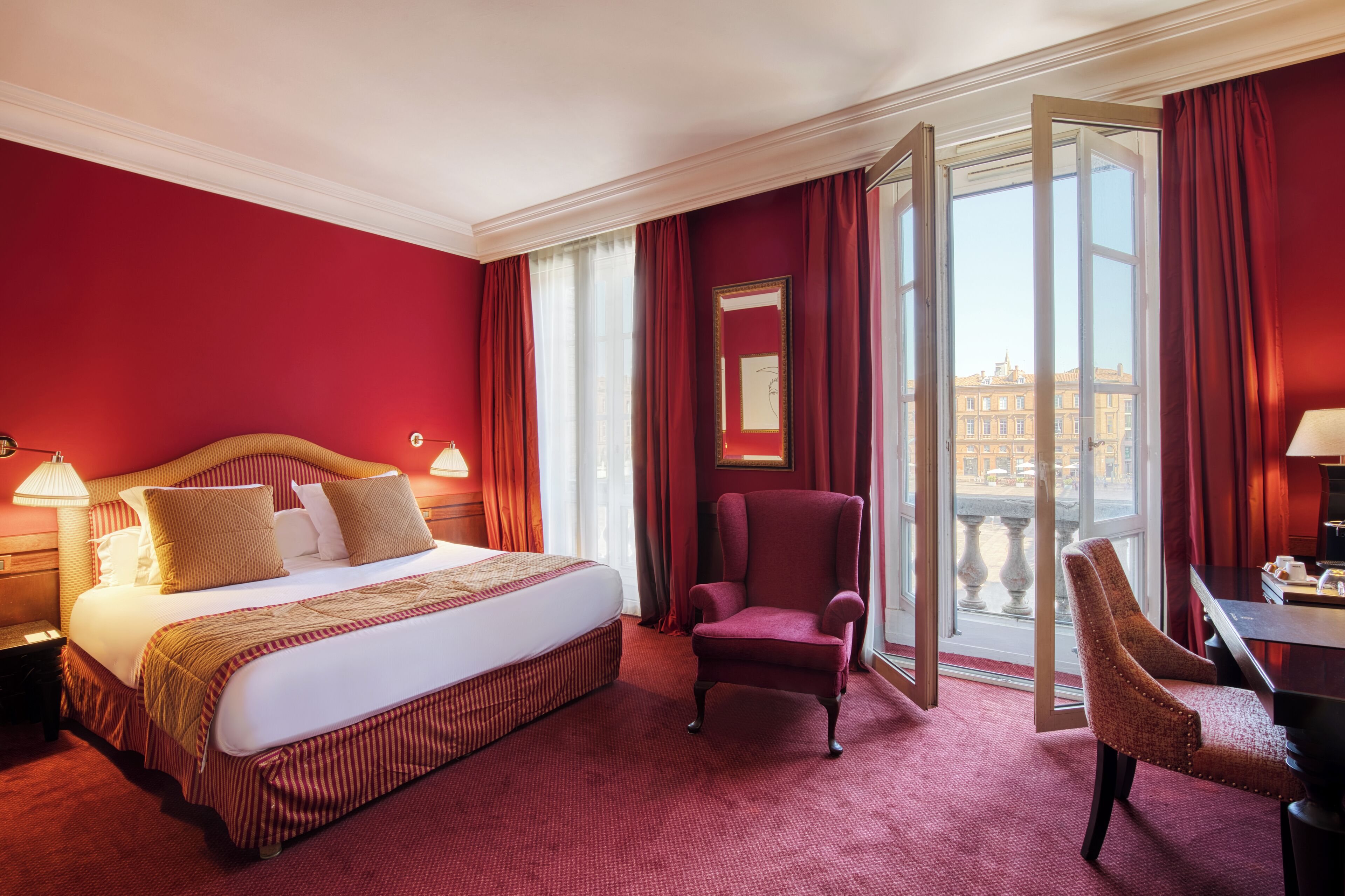 A hotel room with a large bed, a red armchair, a desk, and a view of the city through large windows.