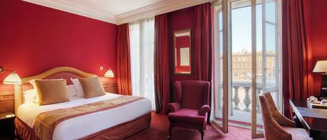 A hotel room with a large bed, a red armchair, a desk, and a view of the city through large windows.