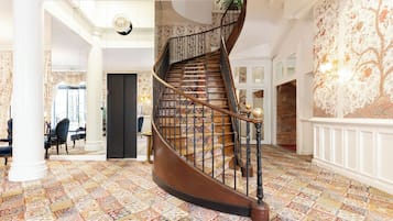 Un magnifique escalier en colimaçon, agrémenté de rampes décoratives et surmonté d’un tapis à motifs, mène à une pièce dotée d’une grande fenêtre et d’un mur recouvert d’un papier peint à motifs floraux.