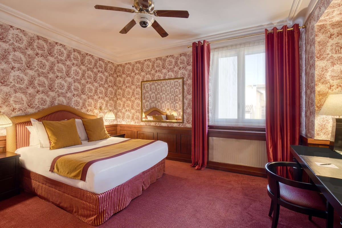 Habitación de hotel con una cama grande, un escritorio, una silla, un ventilador de techo, una ventana con cortinas rojas y un papel tapiz estampado.