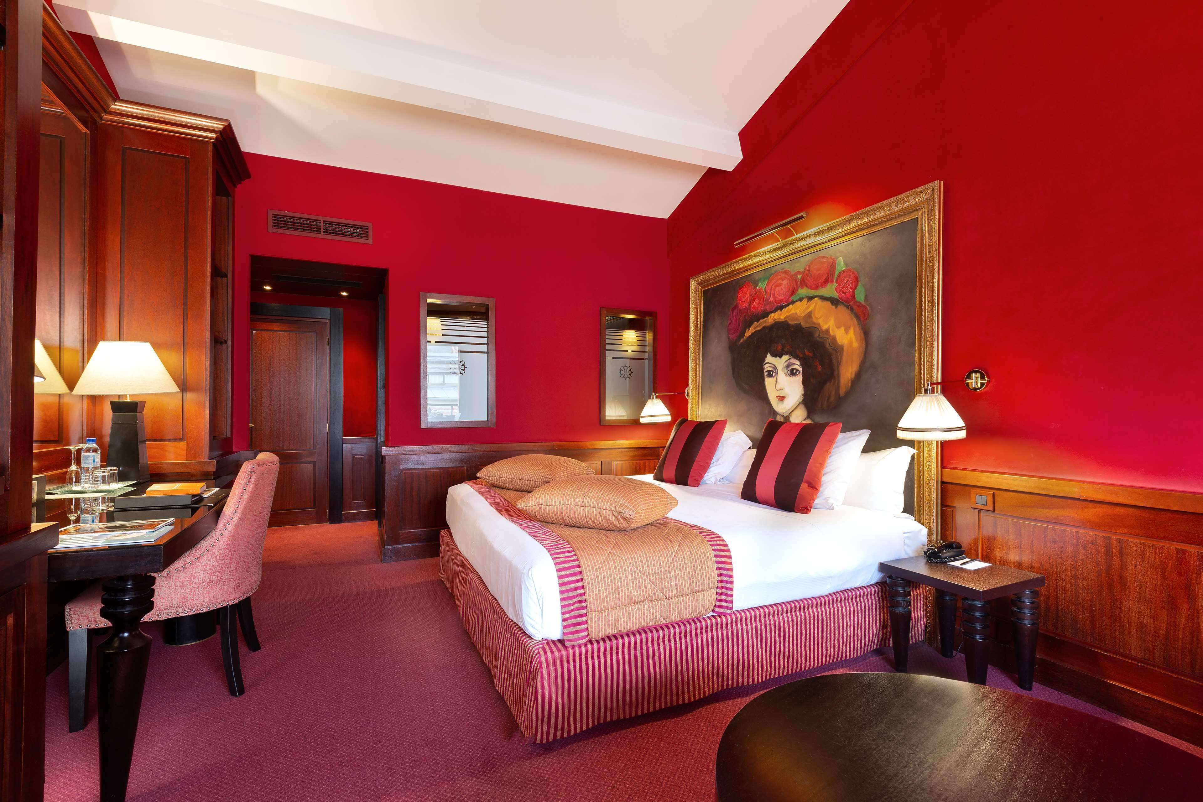 A hotel room with a red wall, a bed with white and striped bedding, a desk with a lamp, and a painting of a woman with a hat.